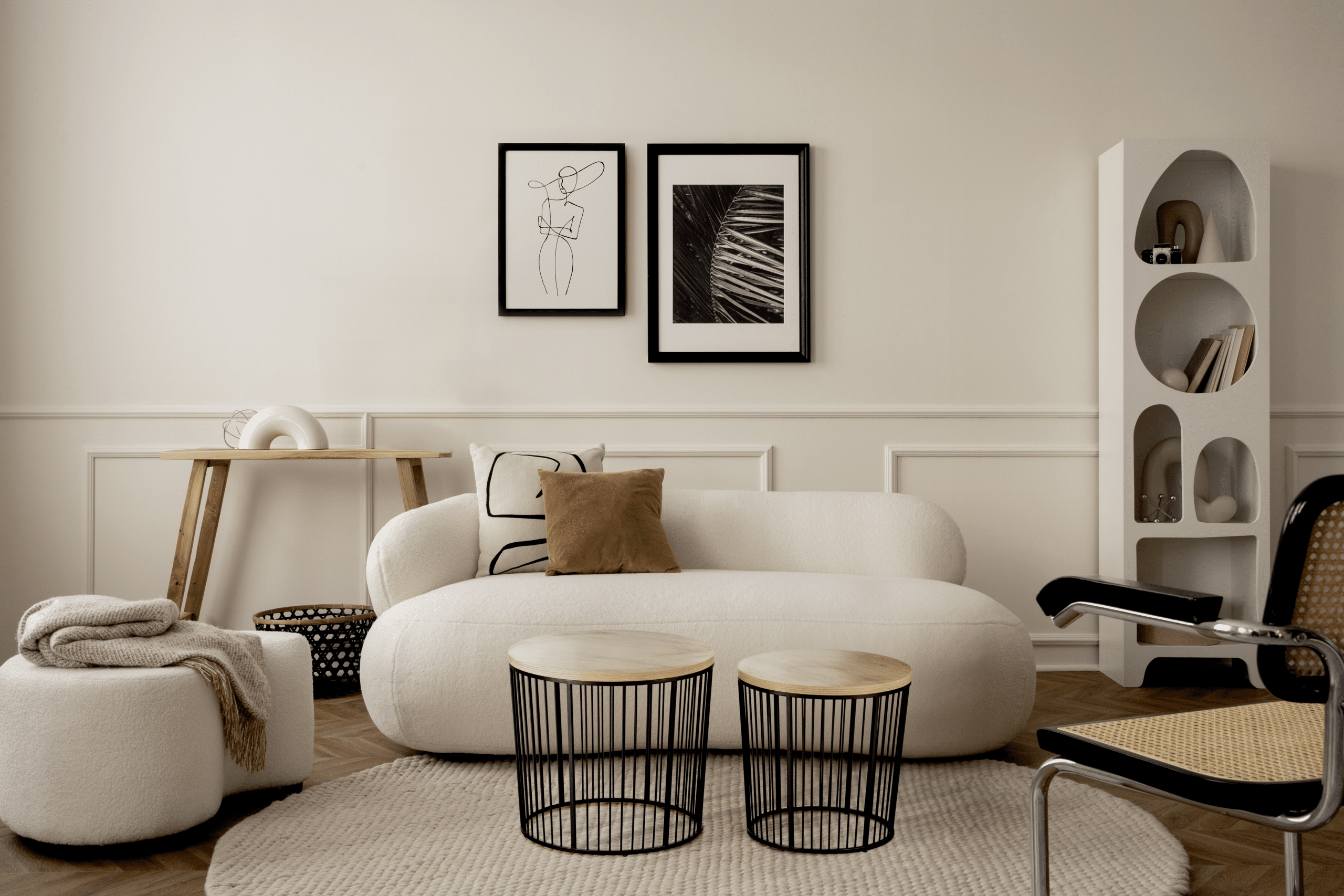 Ein klassisch-modernes Wohnzimmer in Cremeweiss/ Beige gehalten mit einem Sofa und passendem Pouf in amorpher, organischer Form, freiliegendem Teppich, Mobiliar, Accessoires und einem Wandstuck bzw. Vertäfelung im Kassettenstil von ALPHAOMEGA.