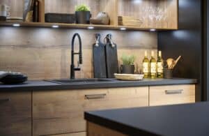 Eine moderne Küche mit anthrazitfarbener Arbeitsfläche und Seitenwänden, offenem Regal, Rückwand und Schubladenfronten im Altholz‑Look und Spotbeleuchtung von ALPHAOMEGA.