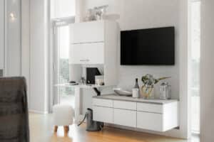 Ein Wohnraum mit Medienboard, klappbarem Hängeschrank in Weiß-Beton‑Look und integriertem Arbeitsplatz von ALPHAOMEGA.
