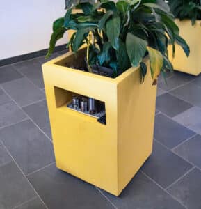 Ein hoher, gelber Blumenkasten mit Ablagefach auf unsichtbaren Rollen von Blumentrog-Flexi.