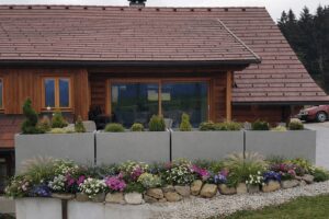 Vier moderne, hohe, winterfeste, graue Pflanzkübel von Blumentrog-Flexi dienen - aufgestellt in L-Form - vor einem Einfamilienhaus als Terrassenabgrenzung.