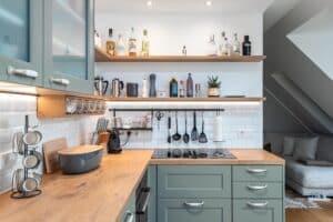 Landhausküche „Classico“ mit Kassettentüren in einem Grün wie Salbei oder Eukalyptus, Oberschränke mit Glaseinsätzen, antike silberne Griffe, Holzarbeitsplatte und gefliester Küchenrückwand mit Utensilienleiste von DANKÜCHEN.