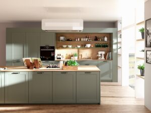 Landhausküche „Lion Classic“ mit Kassettentüren in Grün, Salbei oder Olive, dunklen antiksilbernen Griffen, Holzarbeitsplatte, eingekoffert verbauter Holzrückwand mit Glasregalböden und Kücheninsel mit modernem weissen Dunstabzug von DANKÜCHEN.