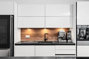 Moderne weiße DAN-Küche mit weissen Hochglanzfronten, Holzrückwand, schwarzer Arbeitsplatte, Spüle und Kaffeemaschine, Modell "Silbermond".