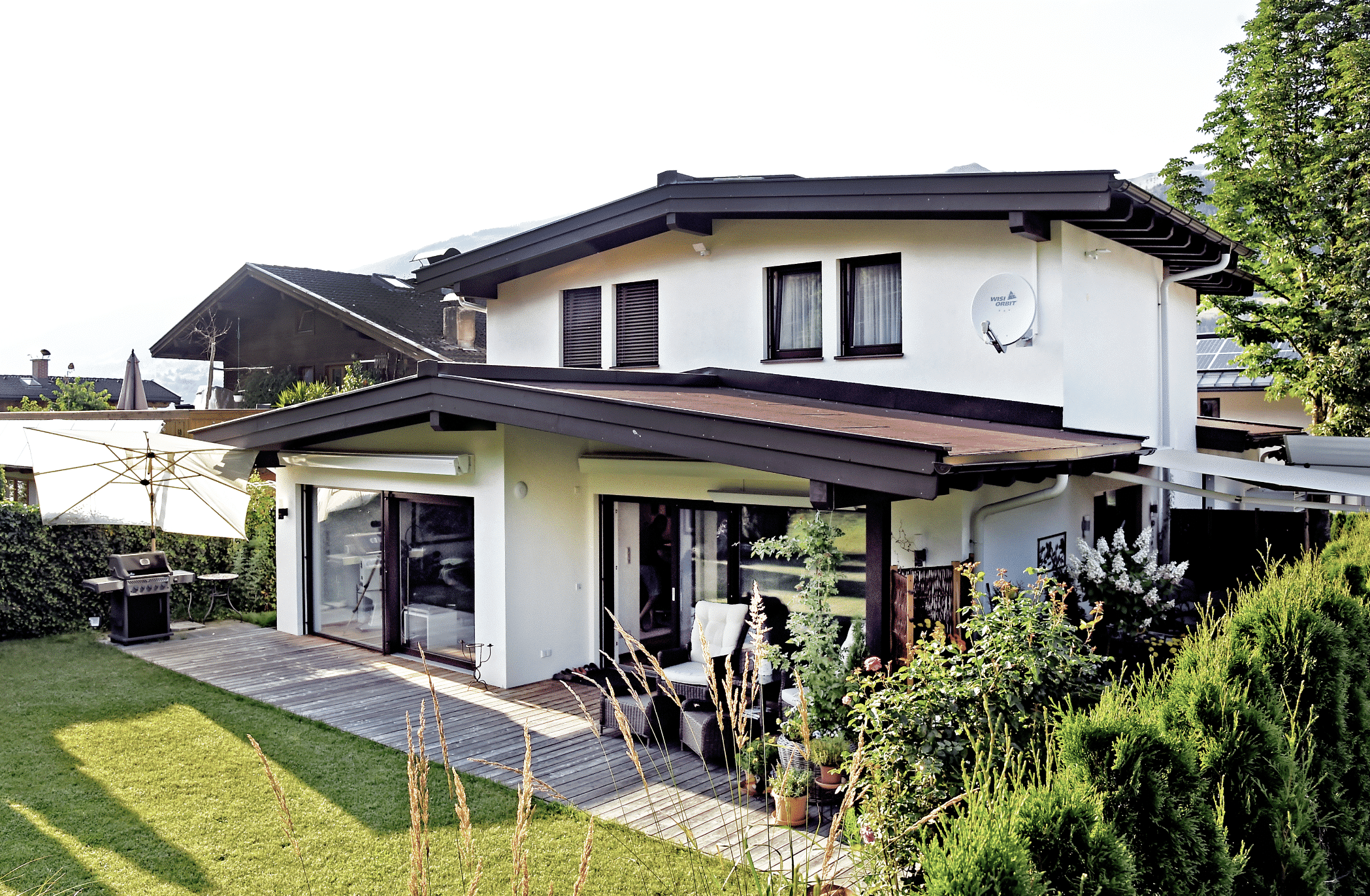 ERLER Haus zeigt ein weisses Einfamilienhaus mit zwei Stockwerken und einer überdachten Terrasse mit Holzboden und einer gemütlichen Sitzecke mit Rattanmöbeln, einem großen Sonnenschirm und einem Grill.