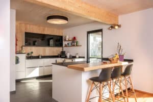 Offene Wohnküche mit Holzvertäfelung an Wand und Decke und einer Inselküche in schwarz-weiss mit integrierter Theke und Barstühlen von ERLER Haus.
