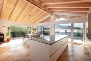 Offene Wohnküche im Obergeschoß eines Hauses mit Holzdecke, balkenmontierter Spotleiste in Schwarz, Panoramaverglasung auf der einen Seite und Wandverglasung mit 2 weissen Schiebetüren zum Eckbalkon und Rauchglasgeländer von ERLER Haus.