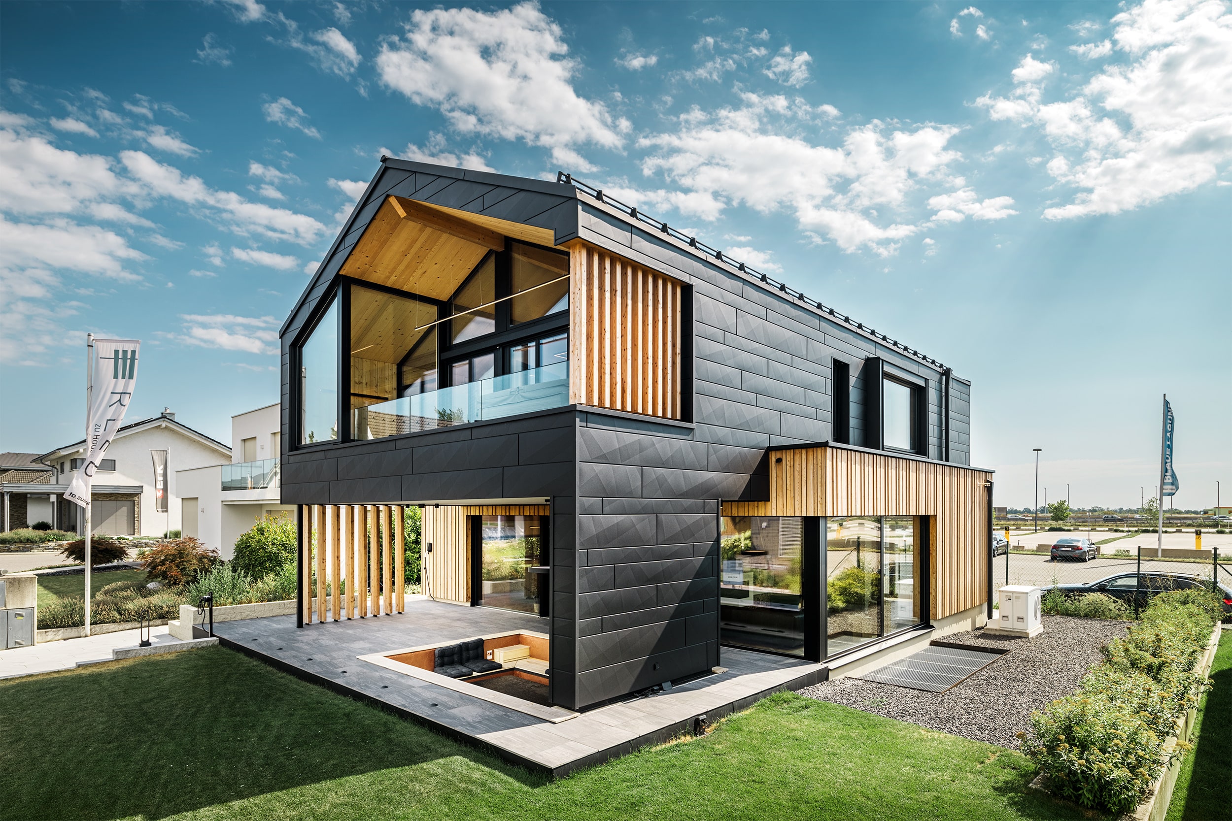 Einfamilienhaus "Autark" mit einem Satteldach mit Solarschindeln, bodentiefen Fenstern und großflächigen Terrassenschiebetüren mit schwarzem Alurahmen, Fassadenelementen aus schwarzem Aluminium, Vollholzsichtwänden, einem Nurglasgeländer in der Loggia des Obergeschosses und einem großem überdachten Terrassenbereich von ERLER Haus.