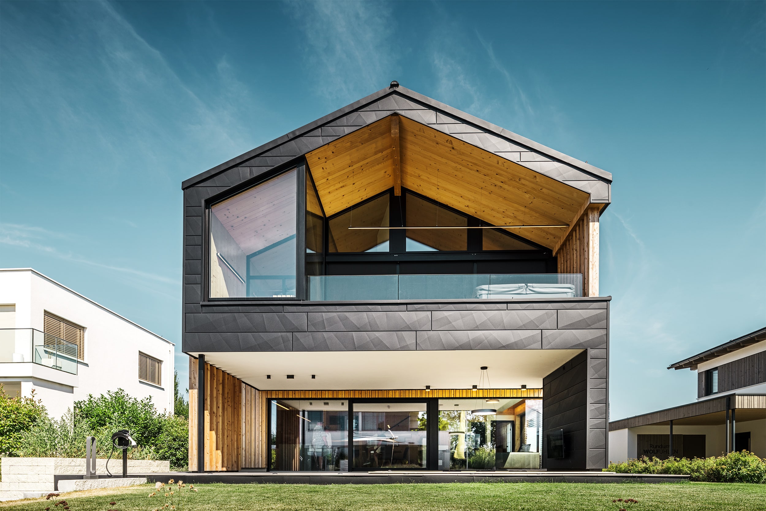 Einfamilienhaus "Autark" mit Satteldach, bodentiefen Fenstern und großflächigen Terrassenschiebetüren mit schwarzem Alurahmen, Fassadenelementen aus schwarzem Aluminium, Vollholzsichtwänden, einem Nurglasgeländer in der Loggia des Obergeschosses und einem großem überdachten Terrassenbereich von ERLER Haus.