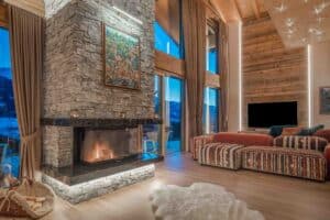 Wonhbereich eines Chalets mit Natursteinkamin, Wandverglasung, Holz-Alu-Terrassenschiebetüren und einer ungetrübten Sicht in die Alpen von ERLER Haus.