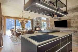 Offene Wohnküche eines Chalets mit Esstisch, Polsterstühlen, rustikalem Natursteinkamin und Terrassenschiebetüren und Verglasung mit Sicht in die Alpen von ERLER Haus.