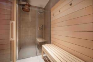 Holzsauna mit Bank und angrenzender, durch eine Glaswand getrennte Duschnische mit Wand- und Bodenfliesen in Steinoptik von ERLER Haus.