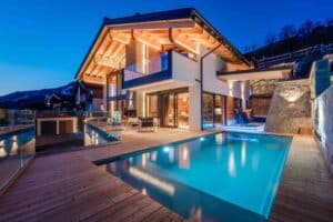 Abendaufnahme einer großzügigen Terrasse eines Chalets mit Holzdielen, Nurglasgeländer, Pool, Jacuzzi, Steinmauer, Outdoormöbeln und beleuchteten Dachbalken von ERLER Haus.