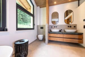 Ein modernes Badezimmer mit raumgetrenntem WC-Bereich, schwebendem Doppelwaschtisch, Holzdecke, Dachschräge, großformatigen Fliesen und großen schwarzen Alu-Fenstern mit Oberlichten von ERLER Haus.