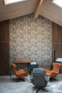Ein einladender hoher Galeriebereich mit drei gemütlichen Loungechairs, schwebenden hölzernen Medienboards, kunstvoller Tapete, Wandvertäfelung aus Holzsprossenelementen, Dachschräge und Dachfenstern von ERLER Haus.