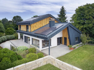 Ein mehrstöckiges Haus in Seitenansicht mit markanter Steinmauer, Holz‑Alu‑Fenstern, Fassadenelementen aus Holz und Blech und dachmontierten Solarpaneelen von ERLER Haus.
