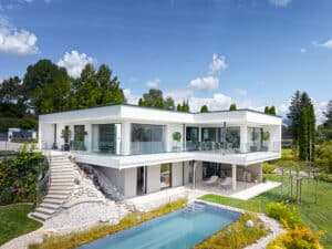 Außenansicht einer modernen Villa mit Flachdach, umlaufendem Balkon mit Nurglasgeländer, Aussenaufgang und einer Terrasse mit einem verspieltem Garten mit Pflasterung und einem in die Natur eingebetteten Pool von ERLER Haus.