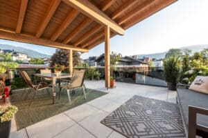 Ein überdachter großer Balkon mit Terrassenmöbeln wie Esstisch und Outdoorcouch, freiliegenden Teppichen und großen Topfpflanzen von ERLER  Haus.