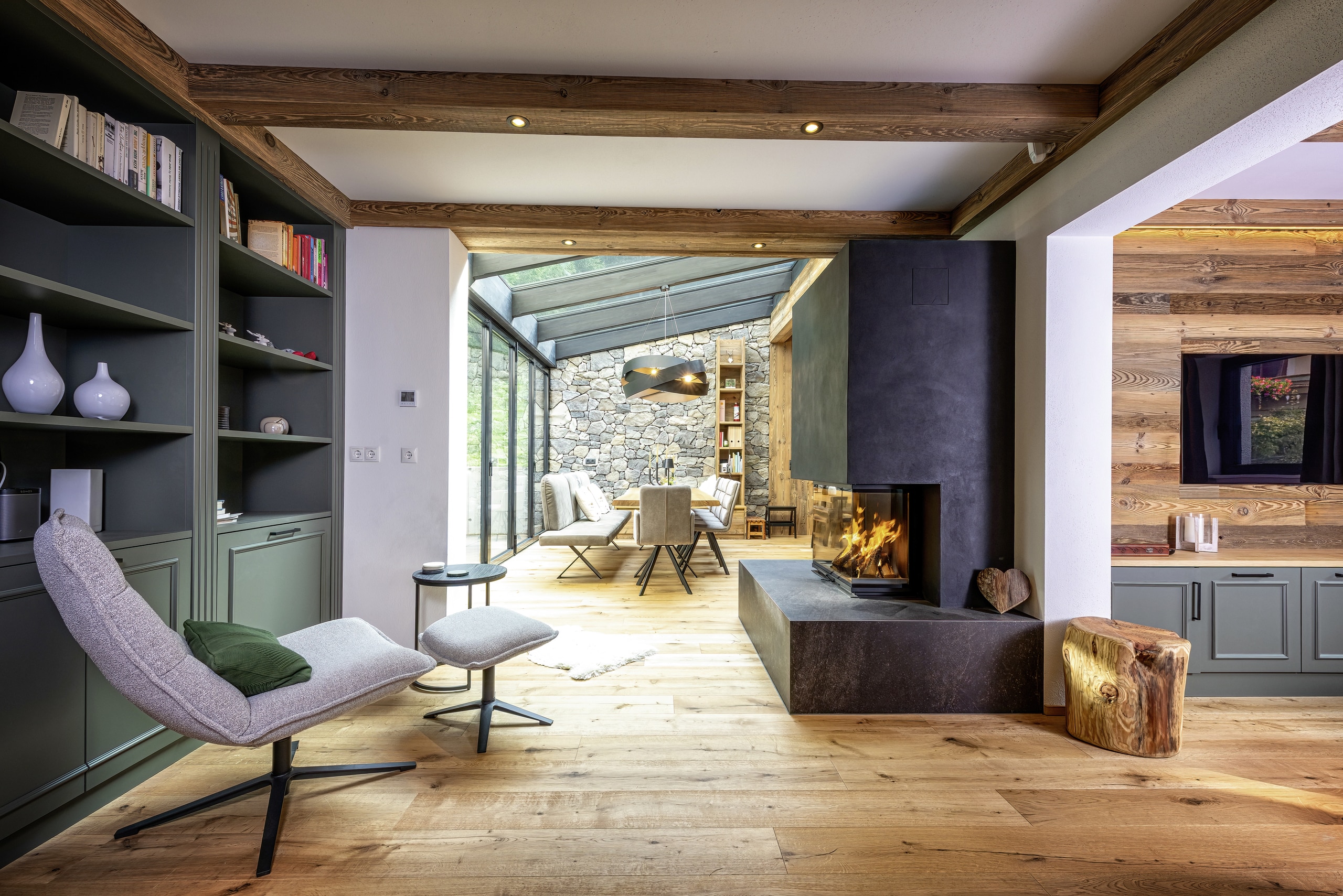 Ein offener Wohnraum mit offenen Nebenräumen, Holzbalken‑Decke mit Spots, gemauertem Kamin, integriertem Bücherregal und angrenzendem Wintergarten von ERLER  Haus.