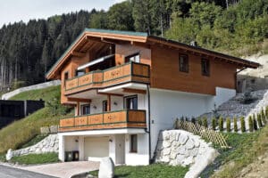 Ein mehrstöckiges Generationen‑ bzw. Mehrfamilienhaus mit rustikaler Holzfassade in Hanglage, mit Garage und zwei überdachten Balkonen mit verspieltem Holzgeländer und Markise von ERLER  Haus.