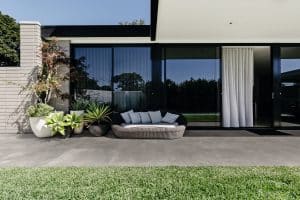 Fliesendorf zeigt eine überdachte Terrasse mit einer gemütlichen Rattanlounge, vielen exotischen Pflanzen und großen Fensterflächen.