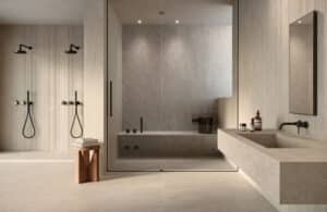 Fliesendorf zeigt ein Wellness-Badezimmer mit großformatigen, fugenlosen Boden- und Wandfliesen in grau-beiger Steinoptik, einem passenden schwebenden Waschtisch, zwei seperate Regenduschen mit Armatur und Stabbrause in Schwarz und eine Steinsauna mit Glastür.