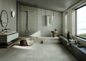 Fliesendorf zeigt ein Badezimmer mit Dachschräge, Bodenfliesen in grau-beige-weisser Steinoptik, einem Doppelwaschtisch und einen erhöhten Aufgang zur Duschnische mit einer Rückwand aus beigen Mosaikfliesen.