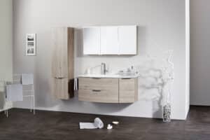 Frauenthal Handel zeigt ein Badezimmer mit einem Waschbecken, Unterschrank aus hellem Holz mit drei Schubladen, schmalen Hochschrank und einem Spiegelschrank mit drei Türen und Beleuchtung.