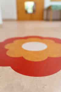 Gerflor zeigt einen beige melierten Linoleum-Bodenbelag mit einer großen orange roten Blume.