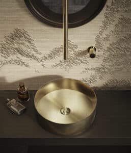 GESSI zeigt ein freistehendes rundes Waschbecken aus Metall mit einer goldfarbenen Armaturen, die an der Rückwand montiert sind und das Wasser über ein langes Rohr mittig auf das Waschbecken trifft.
