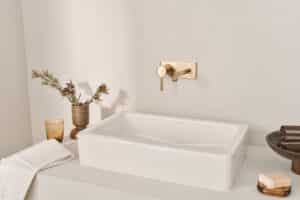 HANSA präsentiert den Wasserhahn mit Drehhebel aus gebürsteter Bronze der Serie Viva, der an der Wand montiert ist, über dem rechteckigen aufgesetzten Waschbecken.