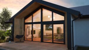 HARTL HAUS zeigt einen eindrucksvollen Bungalow mit Zwerchdach, Wandverglasung mit Terrassenschiebetüren und schwarzem Alurahmen, Fassadenelementen aus Holz und einem Terrassenpodest mit Holzdielen.
