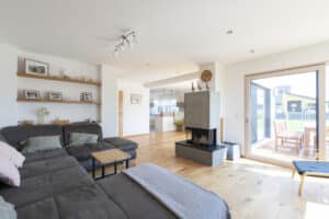HARTL HAUS zeigt einen offenen Wohnraum mit Parkettboden, Sofalandschaft, Kamin und Blick ins Freie und auf die Küchennische.