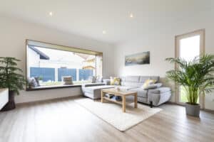 Wohnzimmer eines Einfamilienhauses mit grauem Sofa, einem gepolsterten Sitzfenster mit Panoramasicht und Holzrahmen, und einem bodentiefen Glasfenster mit Holzrahmen von HARTL HAUS.