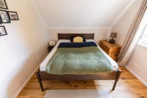 Ein Gästezimmer mit Dachschräge Doppelbett aus dunklem Holz, schwarzem Nachttisch, einer alten Holztruhe und beigefarbenen Wänden für ein Wohlfühlambiente von HARTL HAUS.