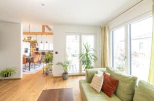 Ein helles Wohnzimmer mit gemütlicher Sitzecke, großem Fenster, stilvollen Holzelementen und offenem Durchgang zur Küche von HARTL HAUS.