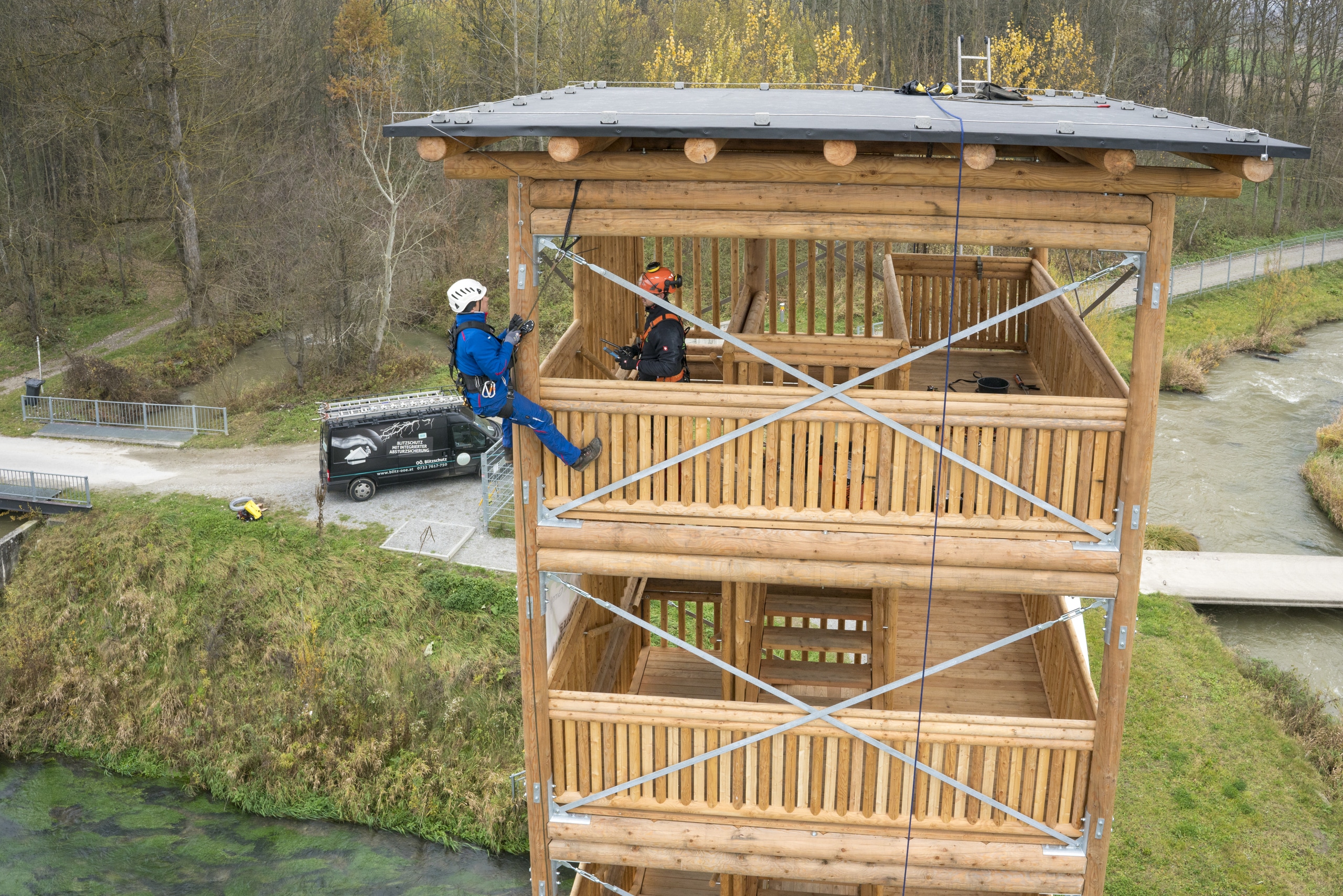 IBS Technisches Büro zeigt zwei Techniker bei der Überprüfung des Blitzschutzes an einem Aussichtsturm über einem Fluss.