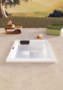 Terrasse mit XL-Steinfliesen und Unterbauversion von Outdoor-Whirlpool "Linos" in Weiss mit schwarzer Nackenstütze bzw. Einstiegshilfe oder Sitzfläche am Rand mit matter Oberfläche und Touchscreen-Panel von Disenia.