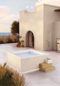 Outdoor-Whirlpool "Gilium" in cremefarbener Ummantelung in Steinoptik mit LED-Beleuchtung am Sockel, Holzstufen und Champagnerkühler auf mediterraner Dachterrasse mit Meerblick, Olivenbäumen und Außendusche von Disenia.