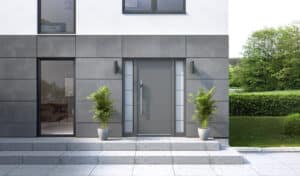 Eine minimalistische Inotherm-Haustür in Anthrazit mit breitem Edelstahl-Stoßgriff und beidseitigen Seitenlichtfeldern aus satiniertem Glas, eingebettet in eine moderne Fassade mit Betonplatten und einer breiten Eingangstreppe mit seitlicher Bepflanzung.