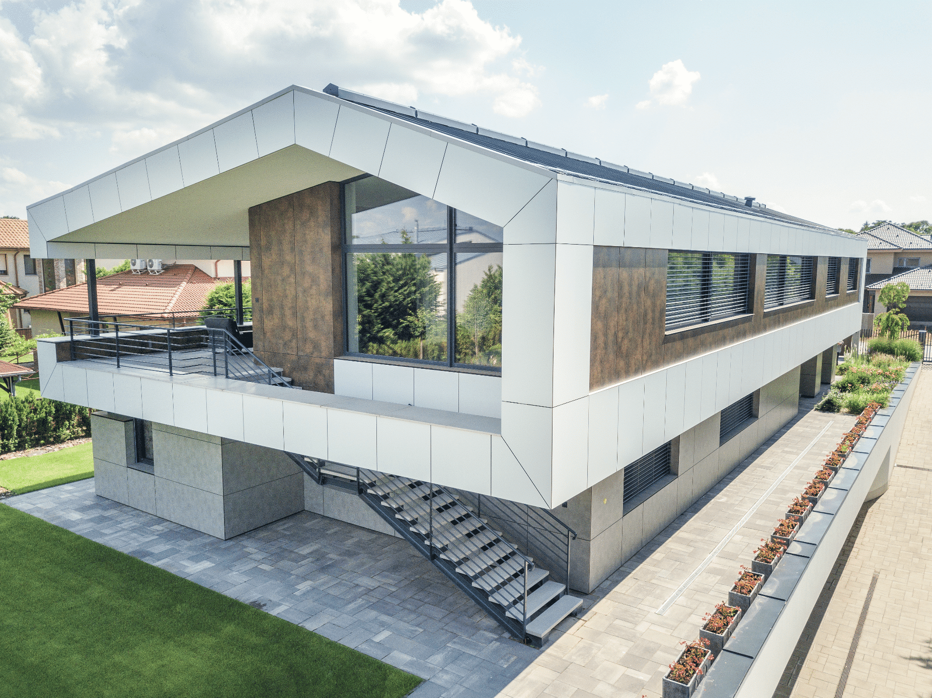 Internorm zeigt ein graues Haus mit weisser Verkleidung beim Dach, großen Fenstern und Steinplatten als Weg um das Haus, mit hellen Treppen und Blumenbeeten.