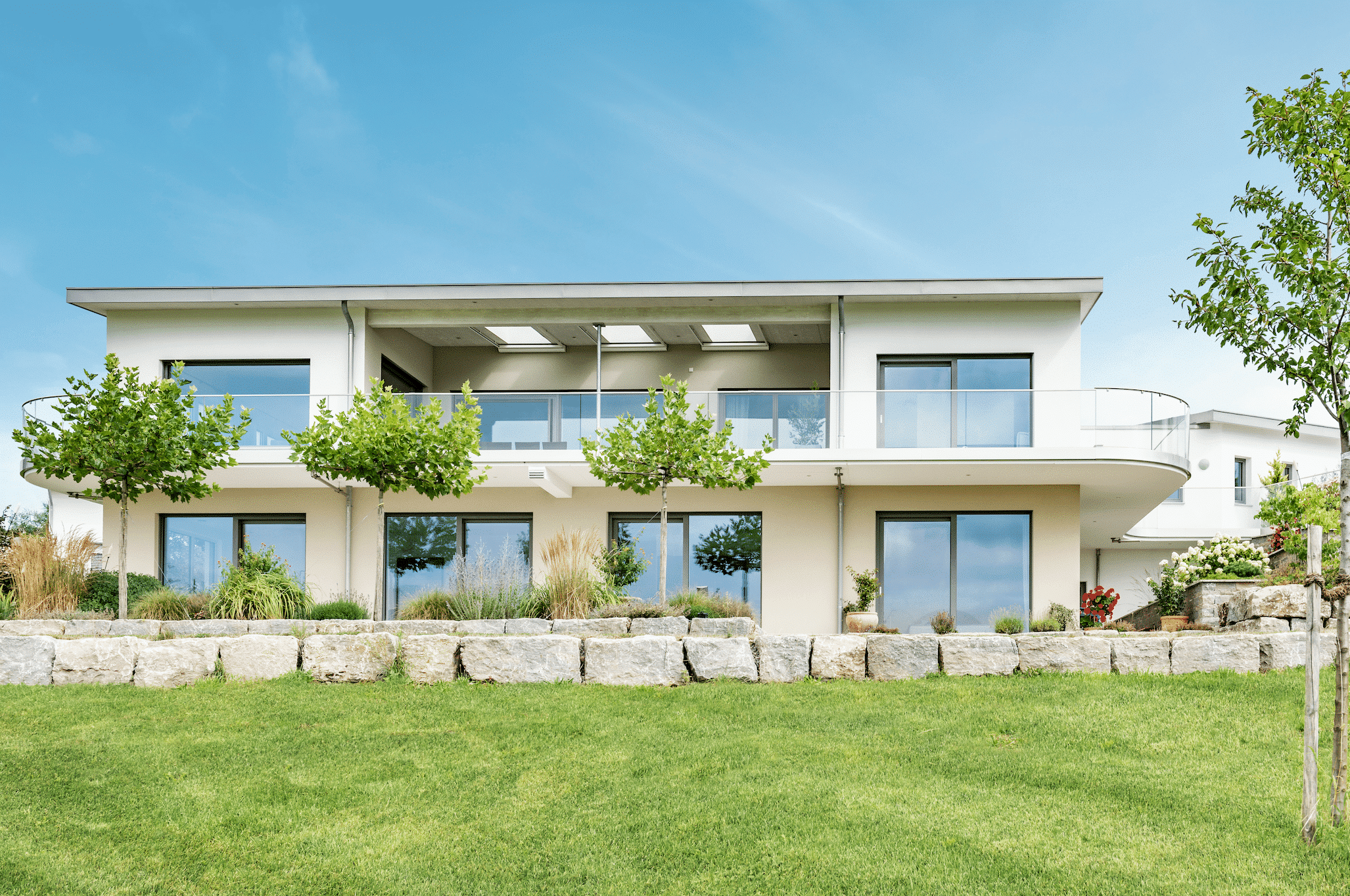 Internorm zeigt ein modernes Haus mit Flachdach und großem Balkon mit Glasgeländer und Glastüren im Erdgeschoß mit Zugang zur Terrasse und dem gepflegten Garten.