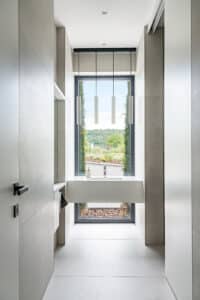 Ein Badezimmer mit schmalem Gang, großformatige Wandverkleidung und Bodenfliesen in Betonoptik und einem bodentiefem Glaselement mit schwarzem Fensterrahmen vor einem schwebenden Waschtisch von Internorm.