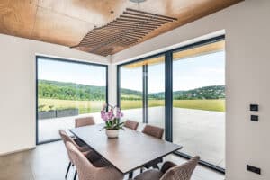 Ein Essbereich mit Wurzelholzdecke, kunstvoller Holz‑Pendelleuchte sowie großflächigen Terrassenschiebetüren und Glaselement mit schwarzem Alurahmen von Internorm.