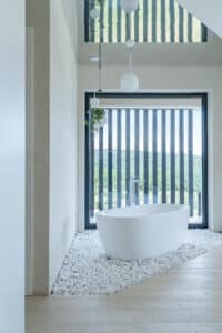 Ein Badezimmer mit Parkett, einer auf weissen Steinen gebetteten freistehenden Badewanne, Spiegeldecke, und einem großen bodentiefen Glaselement mit schwarzem Alu-Rahmen und dazu passenden vertikalen Sprossenelementen im Aussenbereich von Internorm.