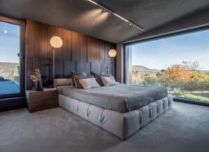 Ein Schlafzimmer mit grauem Teppichboden, passenden Wänden und Dachschräge, dunklem Holzverkleidungselement, einem bodentiefen Panoramafenster und einer Dachbalkontür beides mit schwarzem Alu-Rahmen von Internorm.