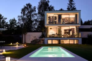 Ein modernes, mehrstöckiges Doppelhaus mit verschachteltem Flachdach, bodentiefen Fenstern, Balkon, Dachterrasse sowie einem Garten mit Pool und Kellerwohnung in Abendbeleuchtung von Josko.