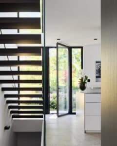 Eine moderne Kragarmtreppe mit Nurglasgeländer im Zwischengeschoß eine schwarz gerahmte Terrassentüre mit Seitenlicht und Bodenfliesen in heller Steinoptik von Josko.