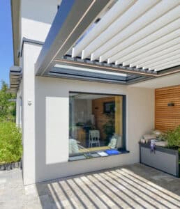 Eine moderne geflieste Terrasse mit Gartenfläche und einer hausmontierten Lamellenpergola mit weißen Lamellen und Gestell in Anthrazit von Moltke.