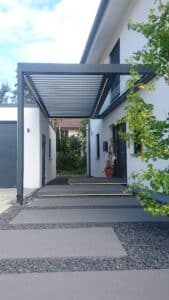 Ein moderner Hauseingang mit einem eleganten - an der Garage endenden - Lamellenvordach in anthrazitfarbener Rahmenkonstruktion, mit integrierter LED-Beleuchtung an Rahmen als auch an den Trittsteinen zum Eingangsbereich - von Moltke.